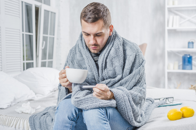 sick-man-sitting-bed-holding-cup-coffee-checking-fever-thermometer_23-2147948517